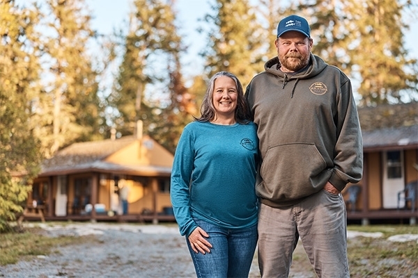 Cindy and Nic, owners of T & D Amisk Camp, standing outside cabins on their property, smiling
